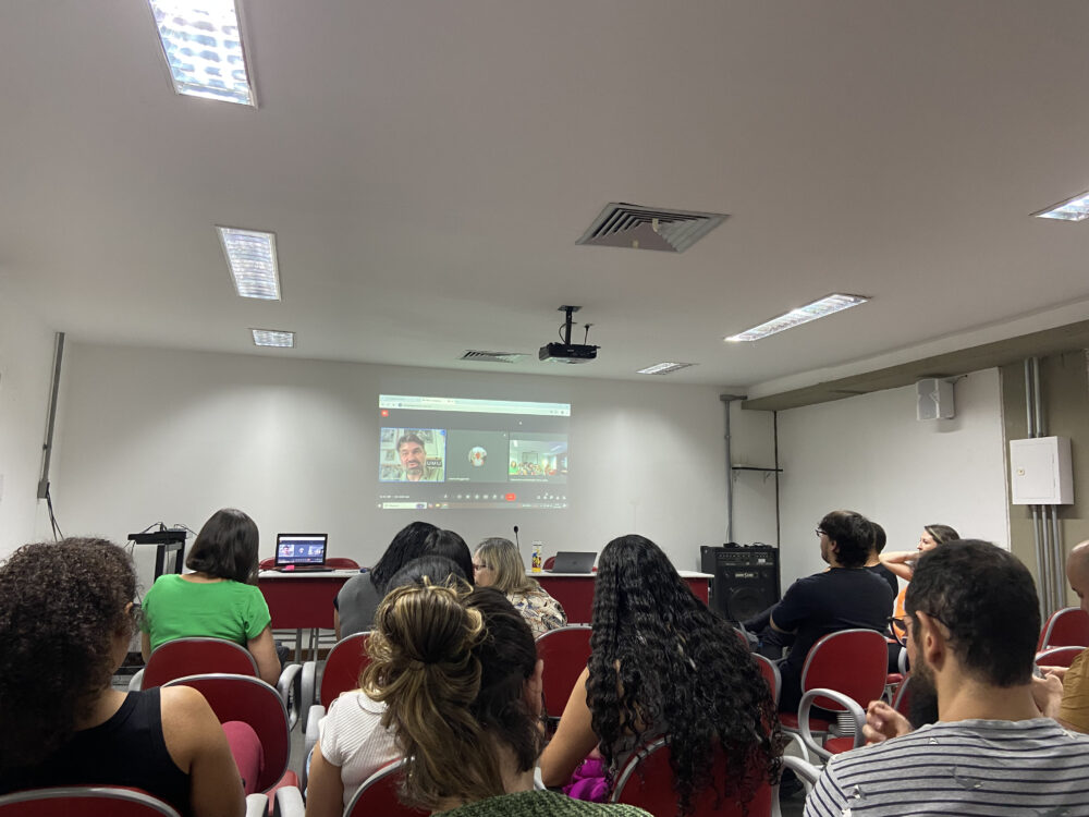 Auditório cheio de alunos sentados assistindo o professor Renato Essenfelder por meio de uma chamada de vídeo projetada na parede